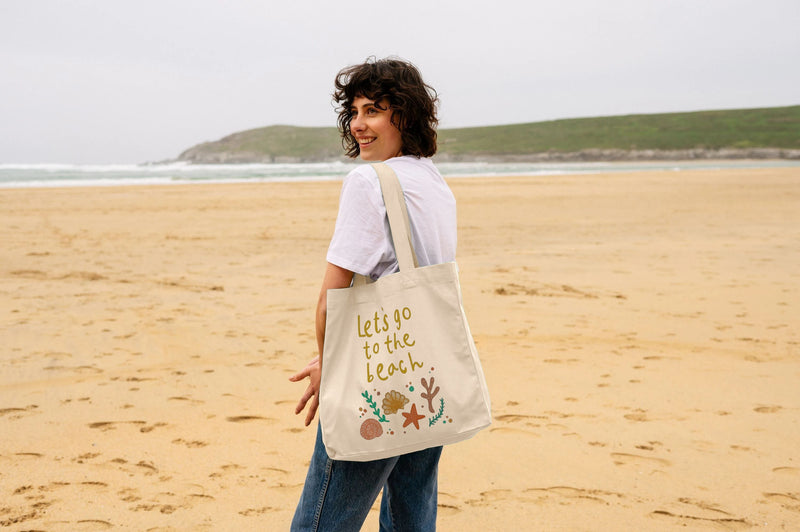 A woman smiling on the beach, carrying a tote bag that says 